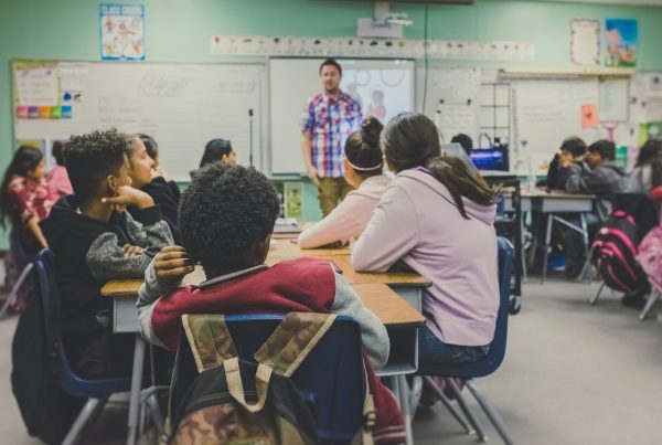 An education setting with a group of students sat in a classroom listening to their teacher, standing in front of the whiteboard